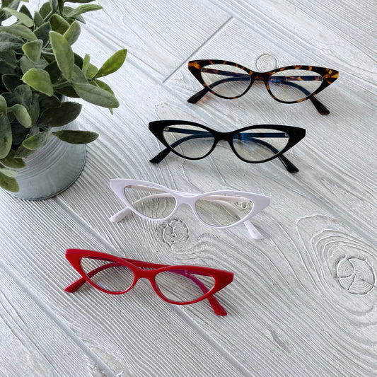 Four pairs of Sophie • Blue Light Glasses in tortoiseshell, black, white, and red are displayed in a row on a light wood surface beside a potted green plant for stylish blue light protection.