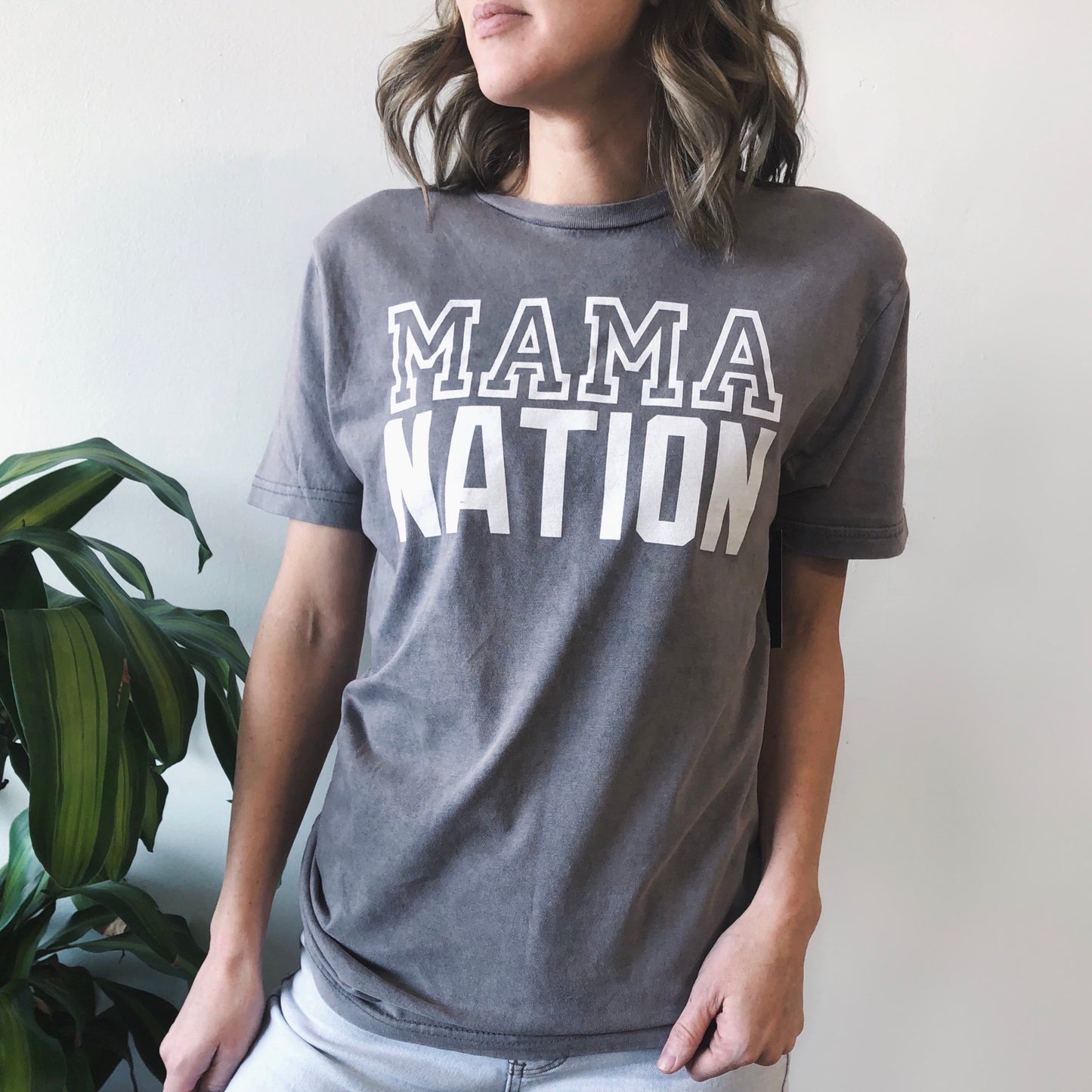 Wearing the Mama Nation • Rocker Tee, a unisex gray shirt with a bold design, a person stands by a leafy green plant against a simple light wall.