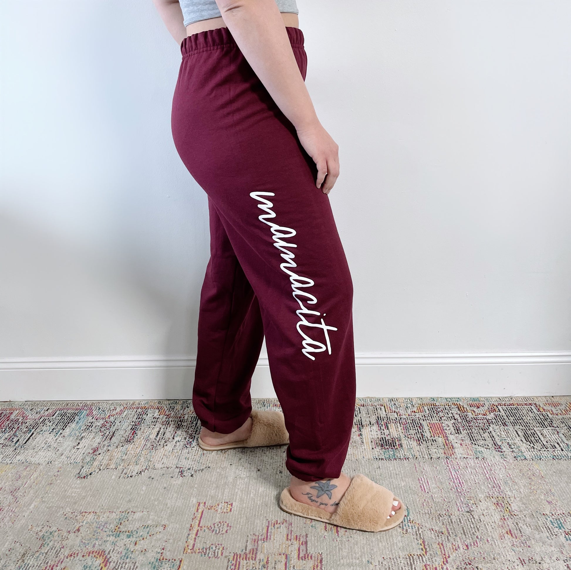 A person stands on a patterned rug against a white wall, wearing Mamacita Sweats • Maroon with “mamacita” in white script, paired with tan slippers and a gray top for a cozy loungewear look.