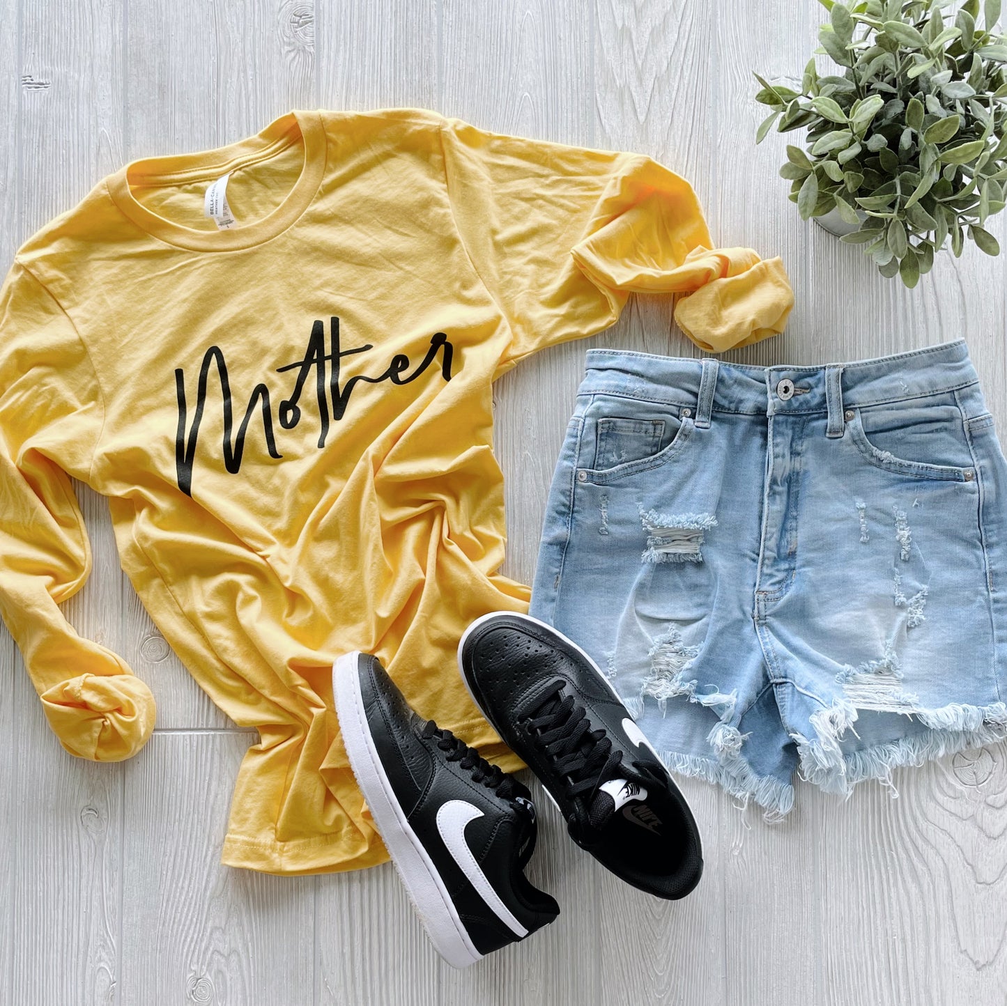 The Mother • Lemon Long Sleeve shirt is styled with light blue ripped denim shorts, black Nike sneakers, and a small potted plant, all arranged on a light wooden surface.