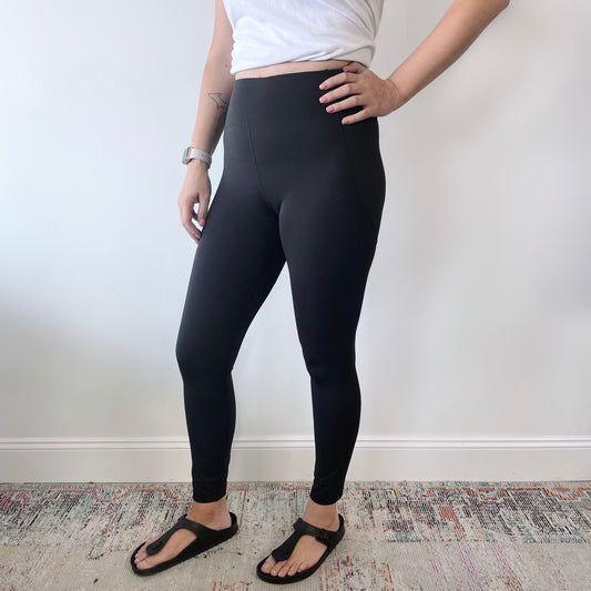 A person stands indoors against a white wall, wearing a white cropped top, the Billie Legging 6209 with tummy control, a smartwatch, and black sandals. Only the lower half of their face is visible.