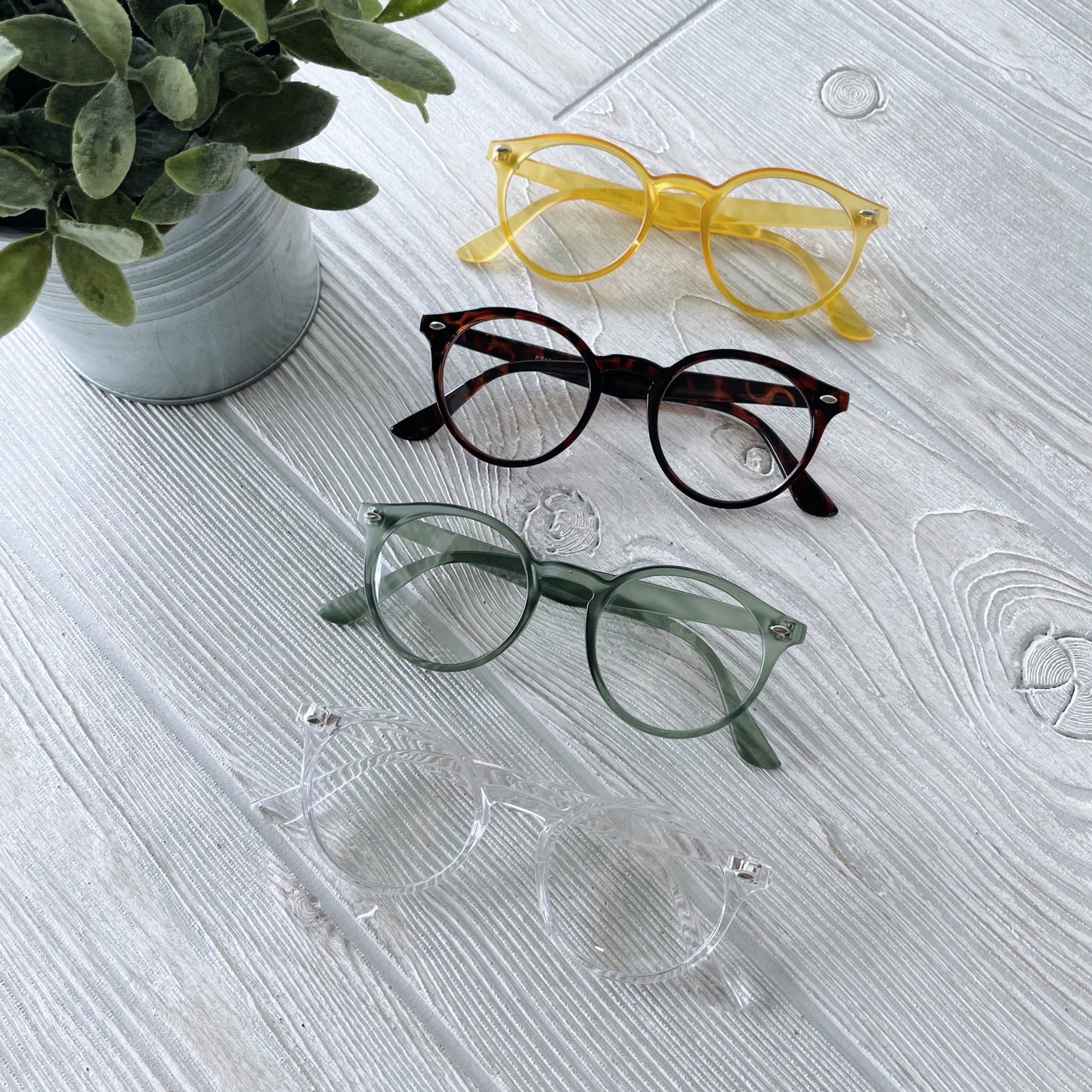 Four pairs of Harper Clear Lens • Adult eyeglasses—yellow, tortoiseshell, green, and clear—are lined up on a white wooden surface near a potted plant. Each pair provides UV400 protection for your eyes.