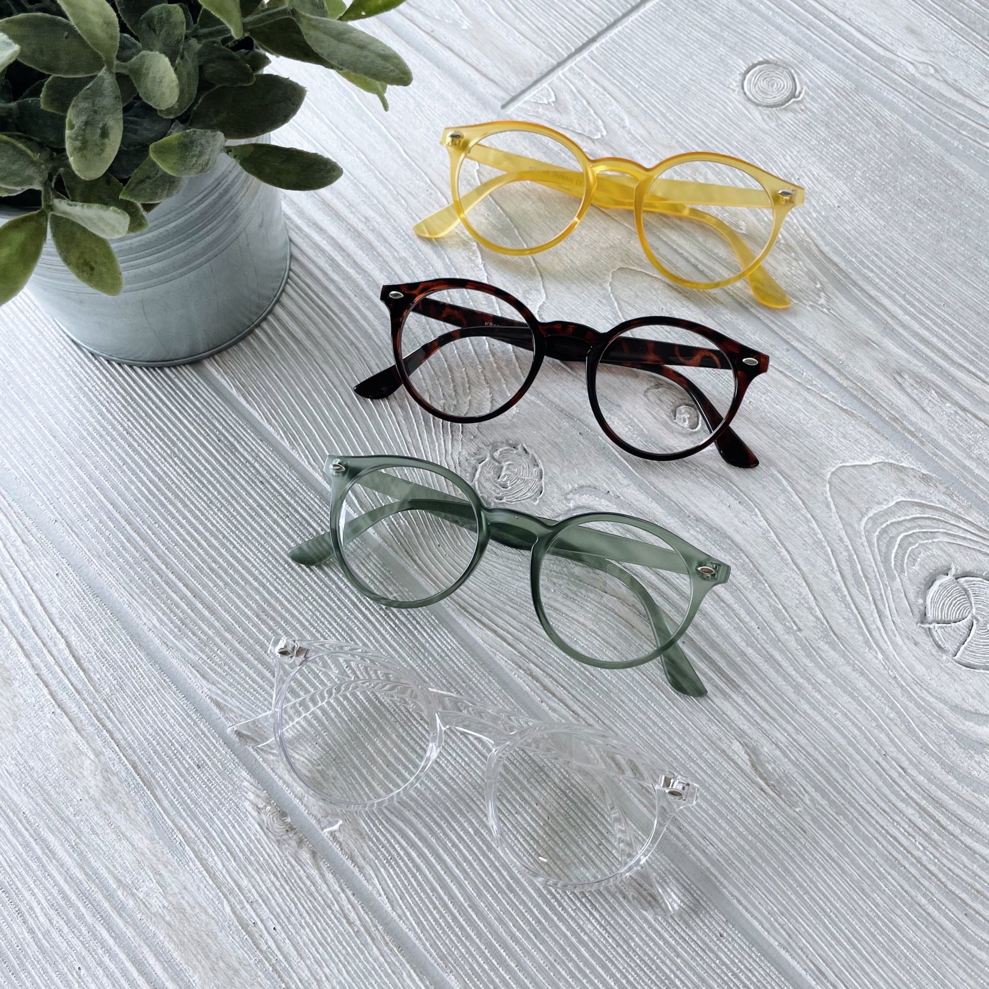 Four pairs of Harper Clear Lens • Adult eyeglasses—yellow, tortoiseshell, green, and clear—are lined up on a white wooden surface near a potted plant. Each pair provides UV400 protection for your eyes.