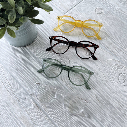 Four pairs of Harper Clear Lens • Adult eyeglasses—yellow, tortoiseshell, green, and clear—are lined up on a white wooden surface near a potted plant. Each pair provides UV400 protection for your eyes.