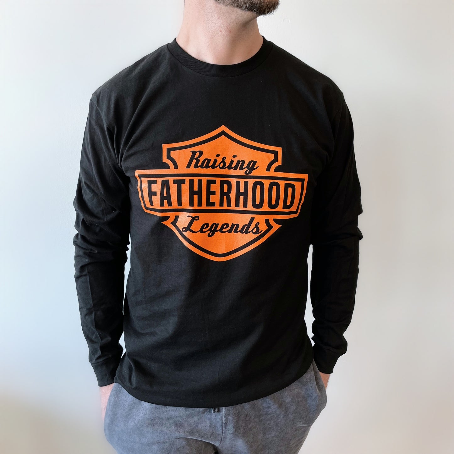 A man wearing the Legends • Dad Long Sleeve—black, unisex, made of soft ring-spun cotton with an orange shield-shaped Raising Fatherhood Legends logo—stands against a plain white background.
