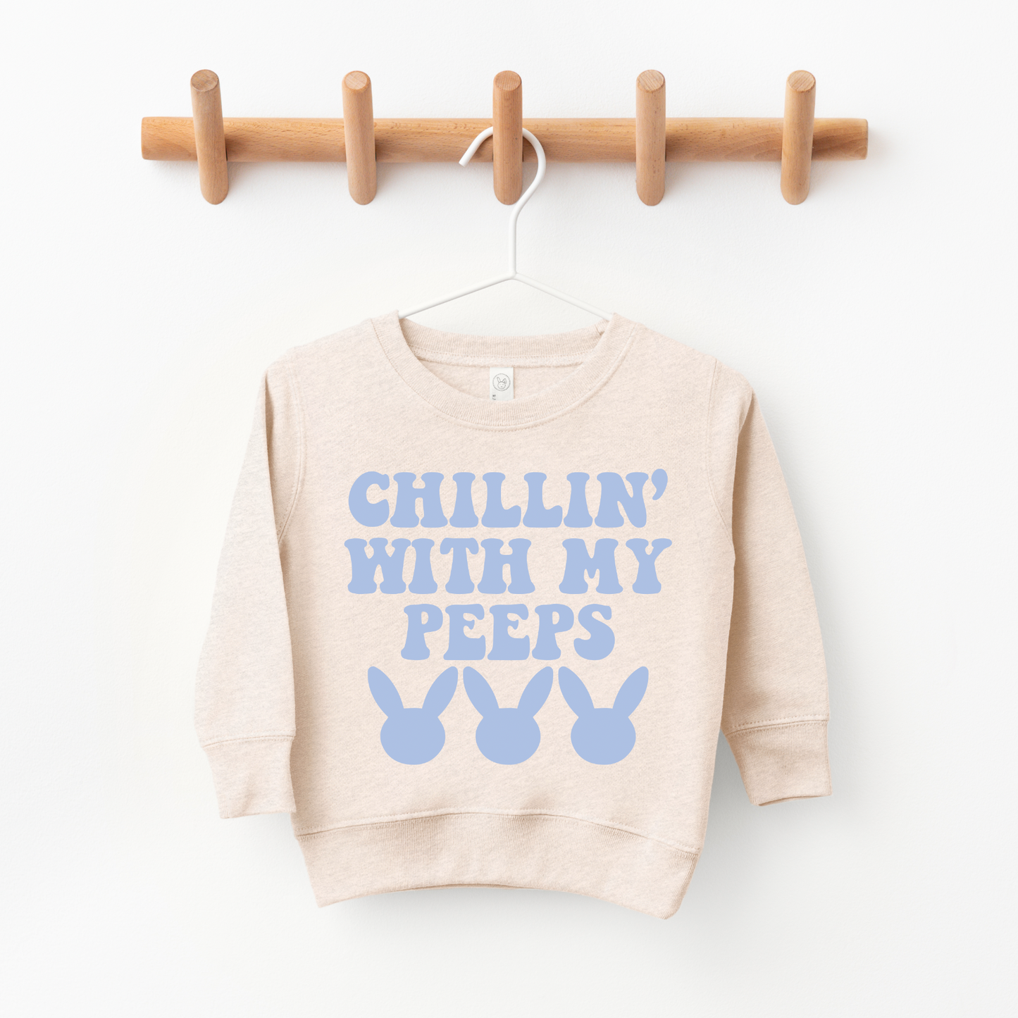 The Chillin Pullover is a beige unisex toddler sweatshirt on a wooden rack, featuring CHILLIN WITH MY PEEPS in blue above three blue bunny head silhouettes—perfect for any kids closet.