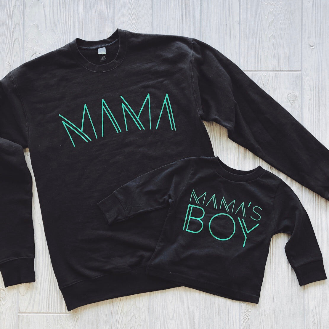 Two black fleece pullovers on a light wood floor: one large with MAMA in lime print, one small with MAMAS BOY. Mama Line Print • Black & Lime Pullover is made from pre-shrunk cotton/poly for a cozy unisex fit.