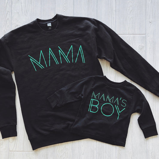 Two black fleece pullovers on a light wood floor: one large with MAMA in lime print, one small with MAMAS BOY. Mama Line Print • Black & Lime Pullover is made from pre-shrunk cotton/poly for a cozy unisex fit.