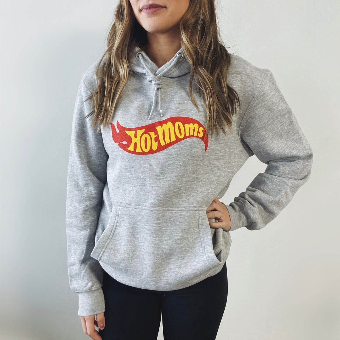 A woman with wavy light brown hair wears the Hot Moms • Gray Hoodie, featuring bold yellow and red lettering, along with black leggings against a plain white background. This unisex hoodie combines cozy comfort and effortless style.