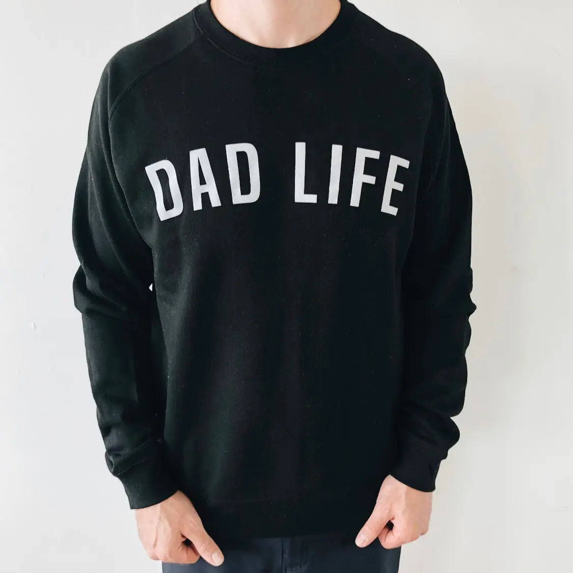 A person wears the Dad Life Block Pullover, a unisex black fleece-lined sweatshirt featuring bold white DAD LIFE text on the front, shown against a plain light-colored wall.