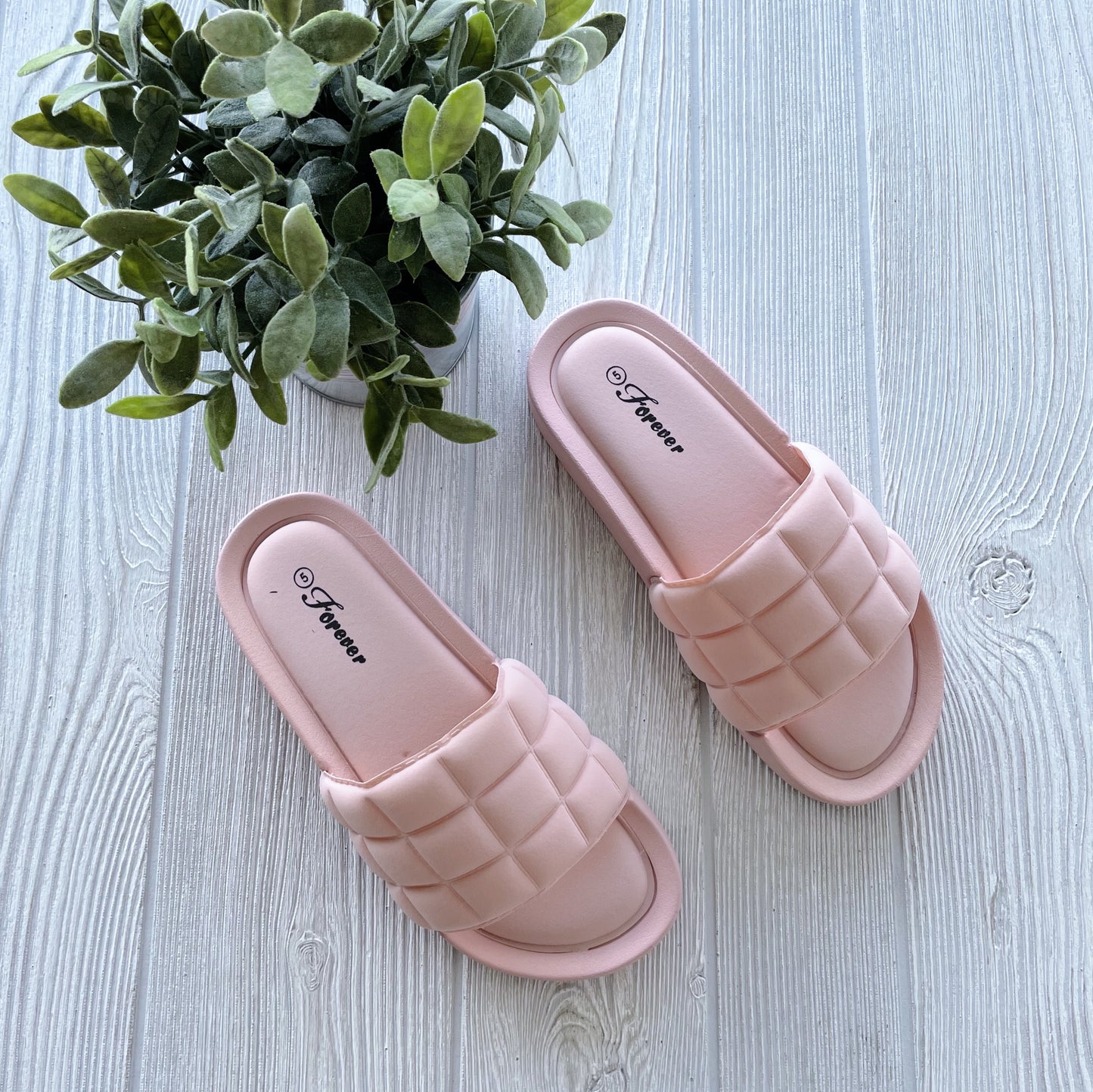 The Halsey Slide Sandal in blush (sizes 5-6.5) features a quilted design and is shown on a light wooden floor beside a green potted plant—ideal for your on-the-go lifestyle.