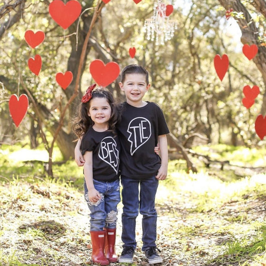 Two smiling children, a girl and a boy, stand arm in arm outdoors wearing matching unisex Bestie • Black Tees. Red heart decorations hang among trees in the sunlit background.