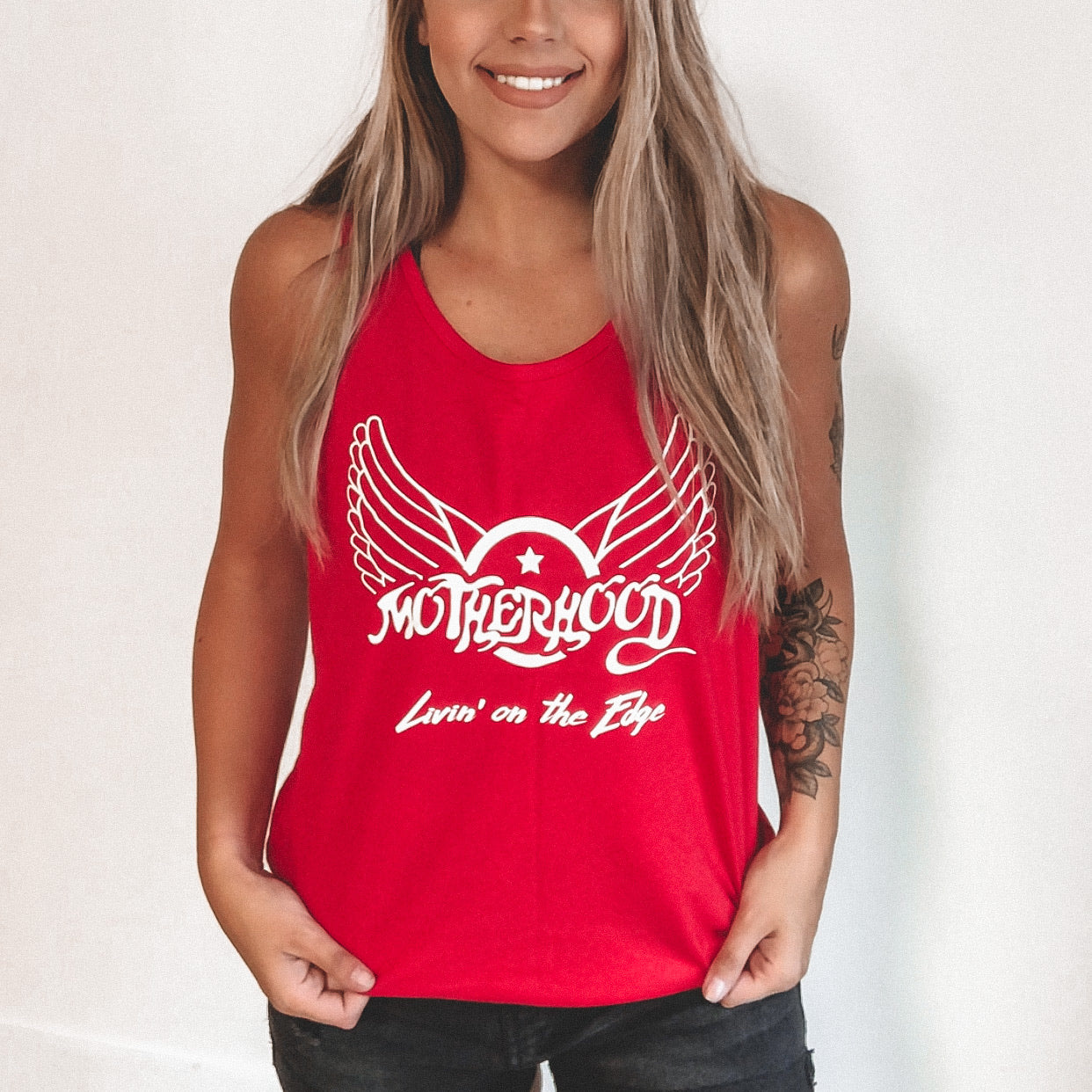 Smiling woman wearing a bright red Aerosmith Mama • Unisex Tank Top with a winged design and the words Motherhood Livin on the Edge, standing against a plain white background.