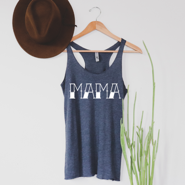 A navy womens tank top with a bold MAMA tattoo print hangs on a wooden hanger beside a brown hat, with a green plant to the right. The background is a plain white wall.