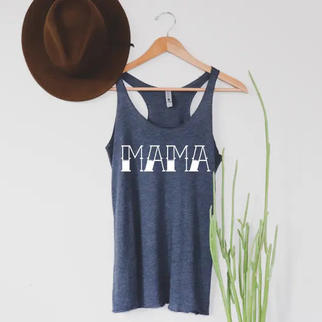 A navy womens tank top with a bold MAMA tattoo print hangs on a wooden hanger beside a brown hat, with a green plant to the right. The background is a plain white wall.