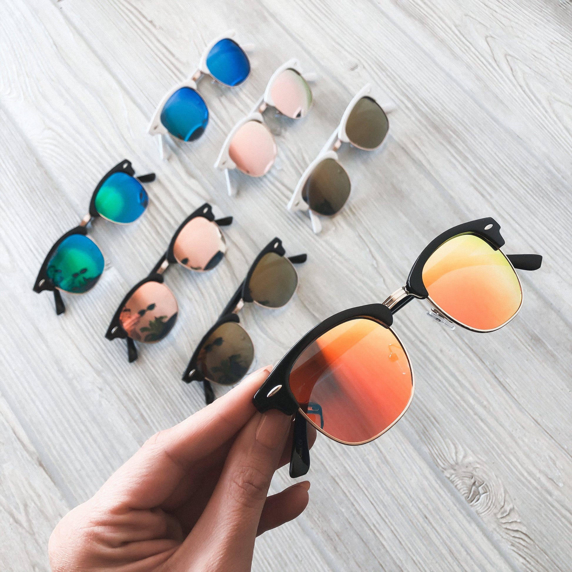 A hand holds Mirror Wayfarer Sunglasses • Adult • More Colors! with orange mirrored lenses and UV400 protection. Six additional adult-sized sunglasses in assorted lens colors are displayed on a light wooden surface nearby.