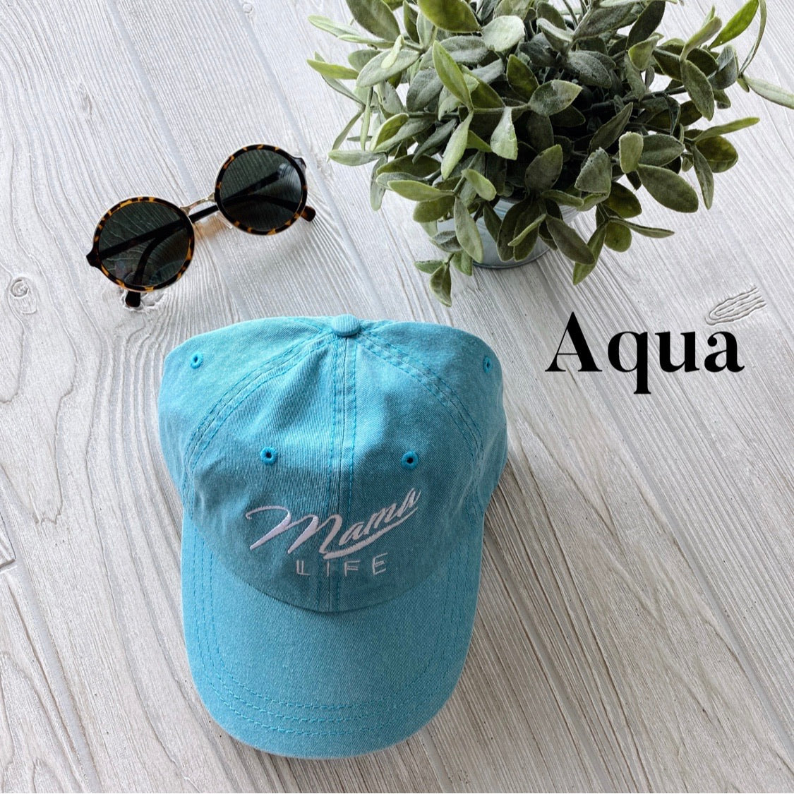 The Mama Life Hat • NEW Colors! in aqua blue is displayed on a light wooden surface, paired with round sunglasses and a potted plant. It features embroidered details and the word Aqua in black text on the right side.