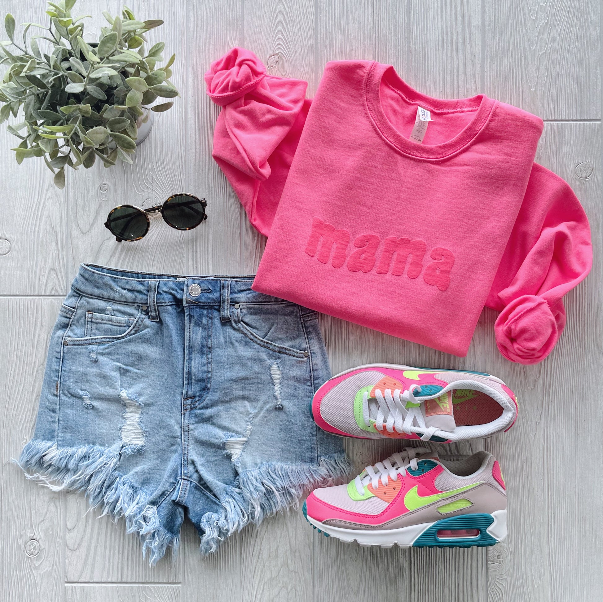 Flat lay of a casual outfit: Mama Wave • Neon Pink Tonal pullover, distressed denim shorts, pink and neon green sneakers, round black sunglasses, and a small potted plant on a light wood background.