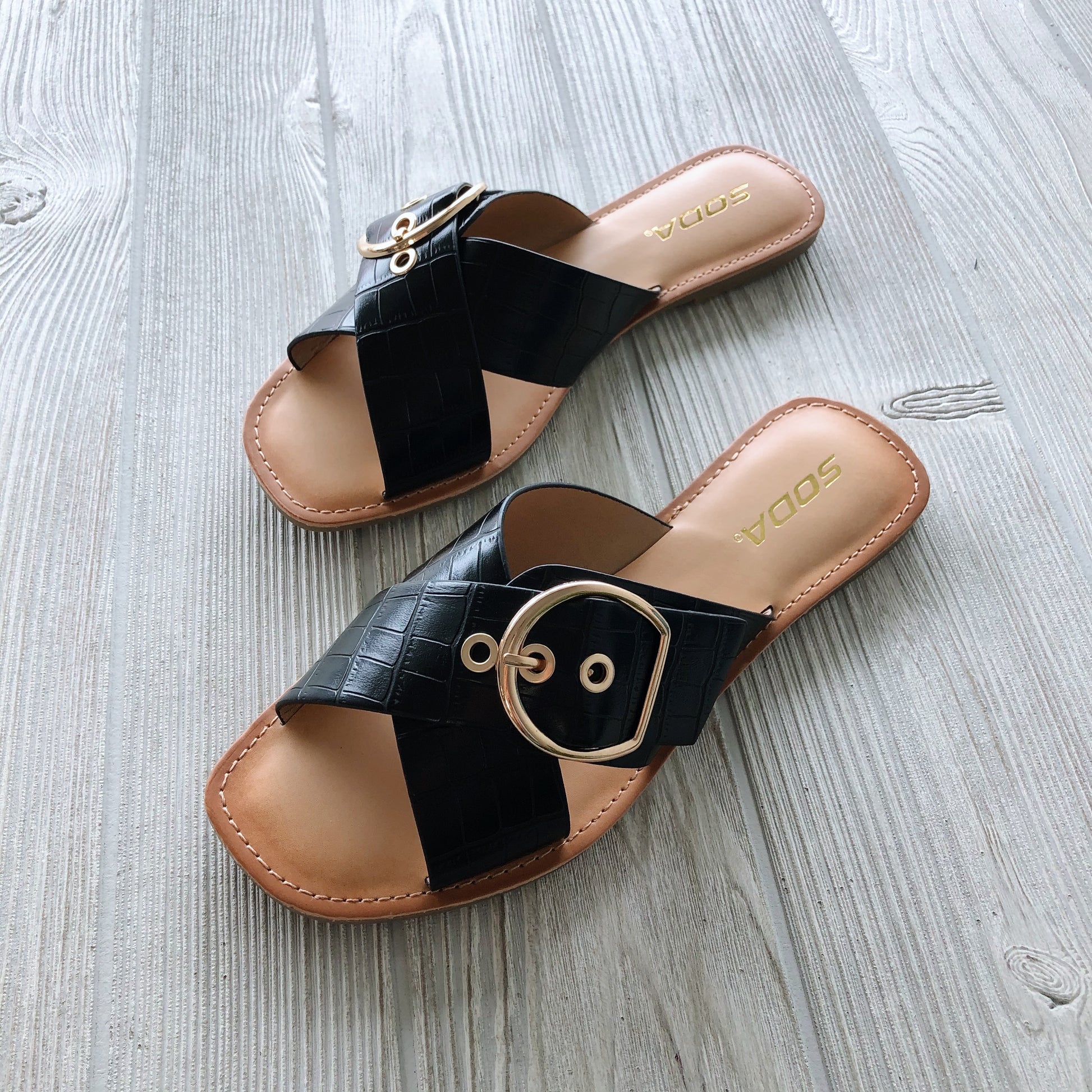 The Buckle Sandal in black features wide crisscross faux crocodile straps, a large gold buckle, tan soles, and the SODA logo on the insole. Shown on a light wood floor. Available in cognac or black for a chic style upgrade!.