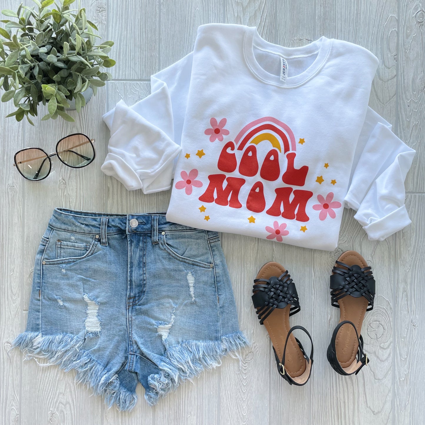 Flat lay of a casual outfit with the COOL MOM • White Pullover, distressed denim shorts, black sandals, sunglasses, and a small green potted plant on a light wood floor.