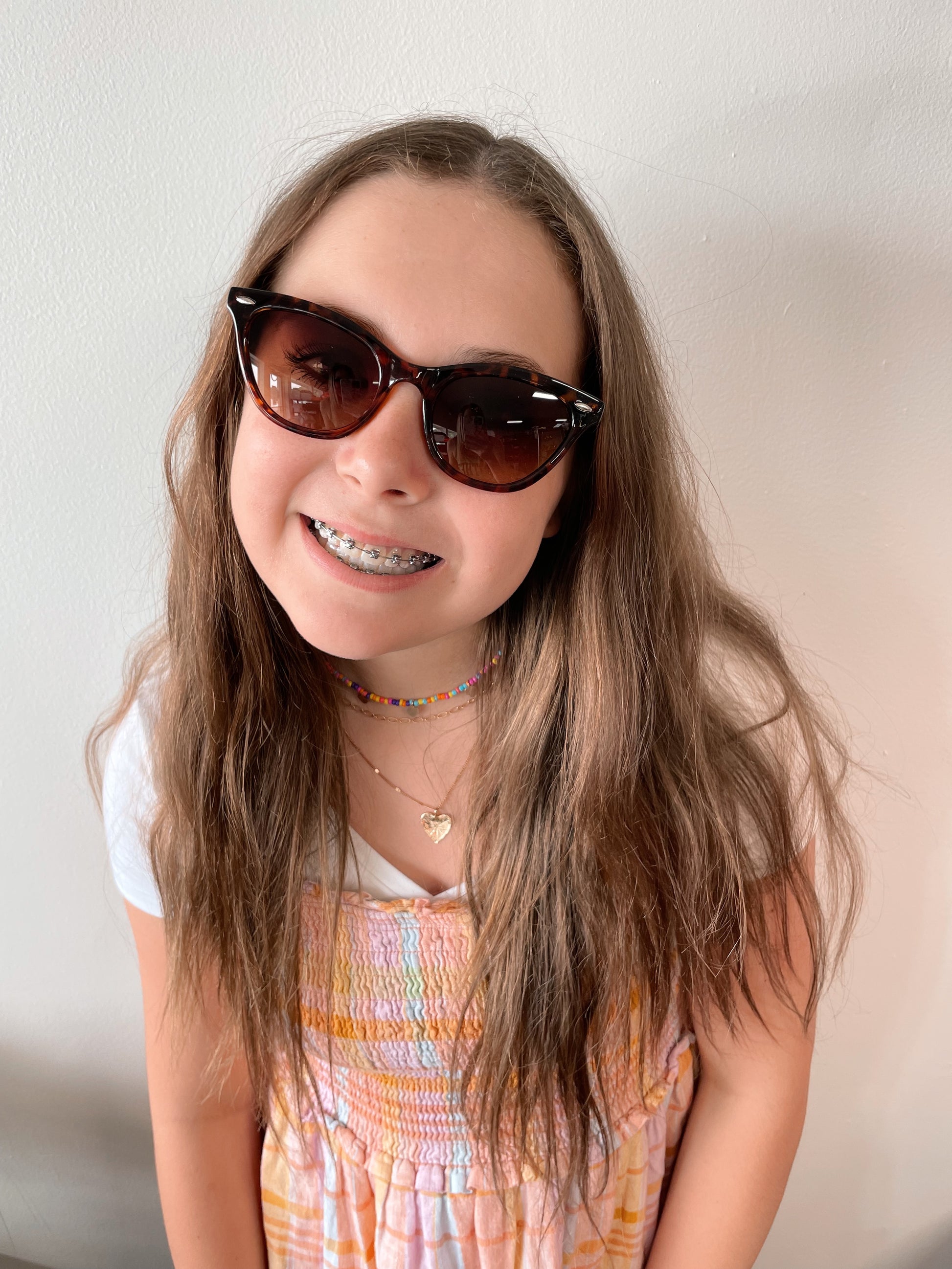A smiling girl with long brown hair, braces, and a colorful beaded necklace wears Bambi Sunglasses • Adult (UV400 protection), a white t-shirt, and a pastel striped dress while standing against a light-colored wall.