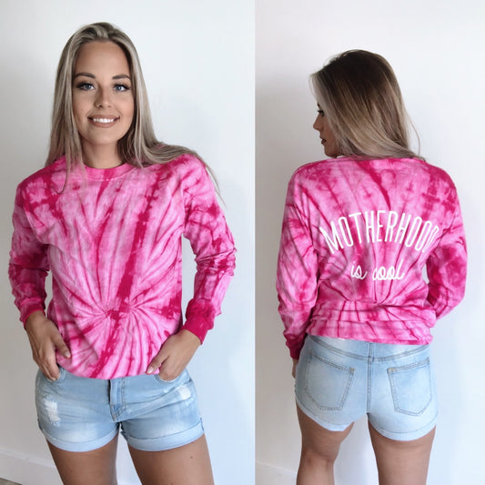A woman models our Motherhood is Cool • Pink Longsleeve Tie-Dye, featuring a bold back design, paired with light denim shorts. She poses both facing forward and showing the “Motherhood is cool” statement on the back.