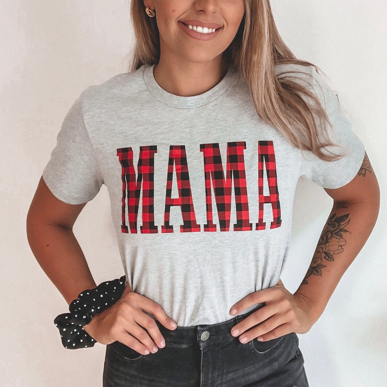 A woman with long hair, a tattoo, and a black polka dot scrunchie smiles with hands on hips, wearing the MAMA • Buffalo Plaid tee featuring a bold buffalo plaid MAMA print.