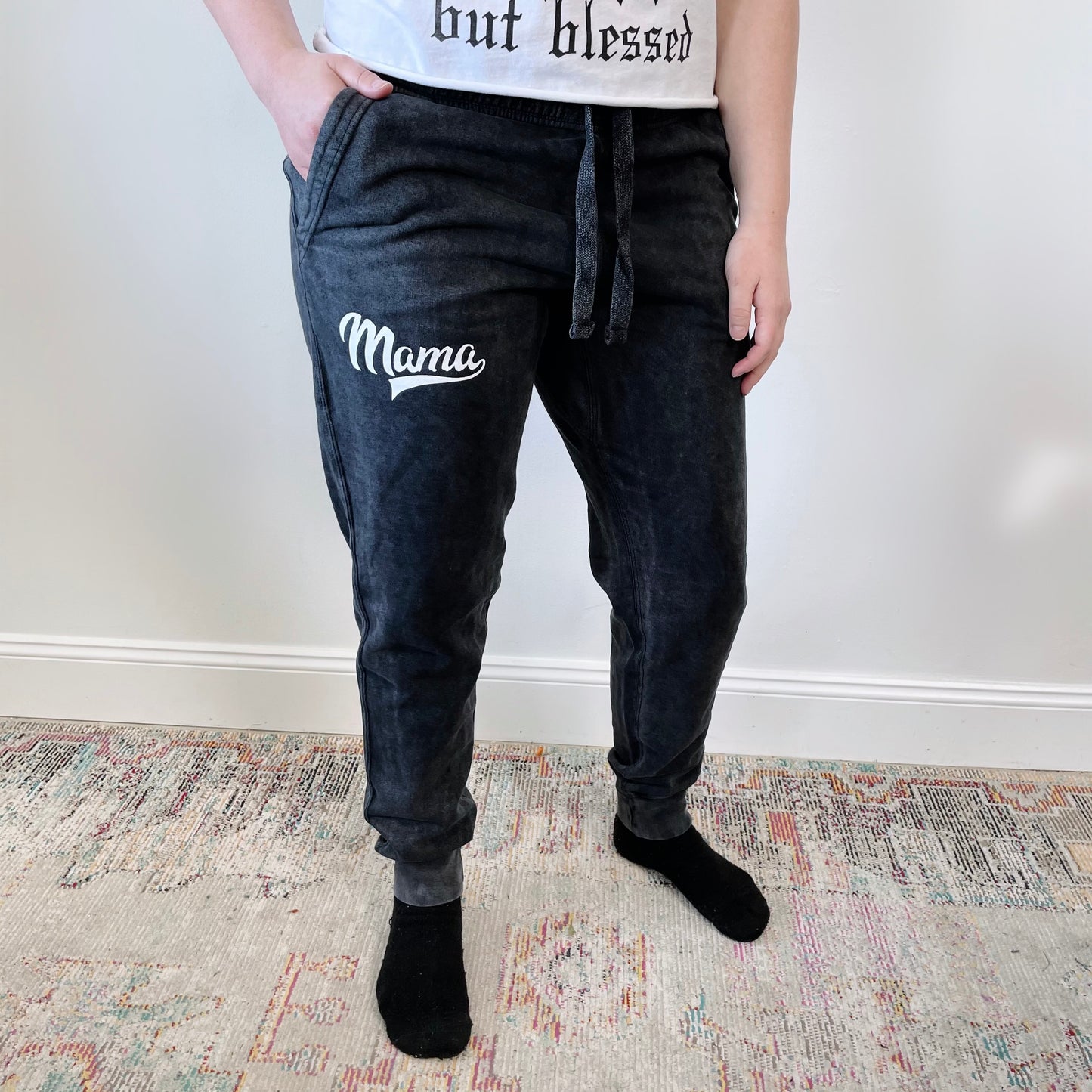 A person stands on a patterned rug against a white wall, wearing Mama Sporty • Acid Wash Joggers, a white shirt, and black socks.