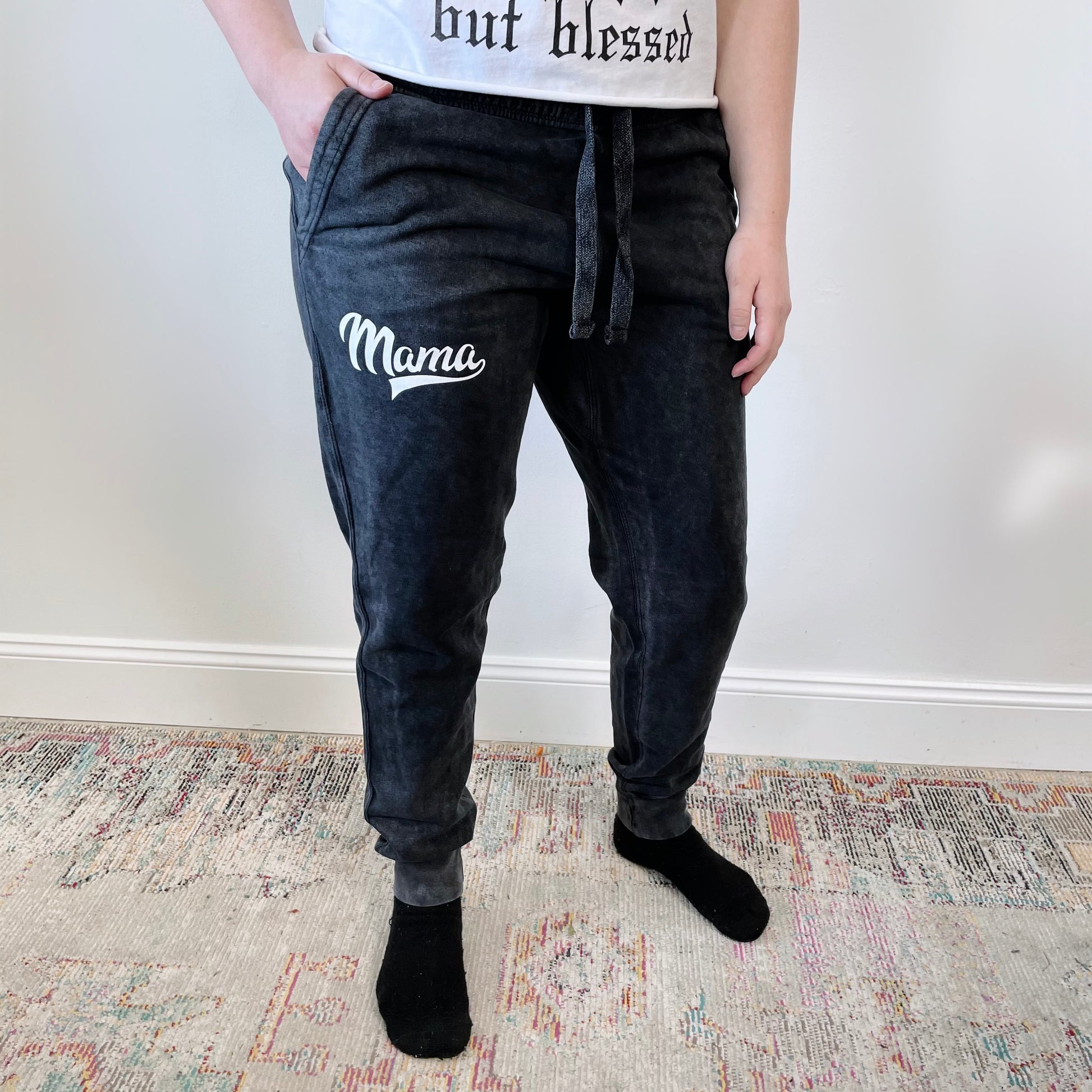 A person stands on a patterned rug against a white wall, wearing Mama Sporty • Acid Wash Joggers, a white shirt, and black socks.