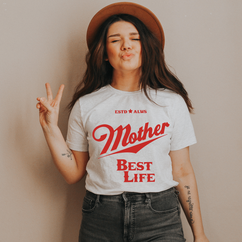 A woman wears a unisex Bella/Canvas Ash Best Life Tee, with Mother Best Life in red text, posing playfully against a beige wall—peace sign up, lips puckered, and wearing a hat.