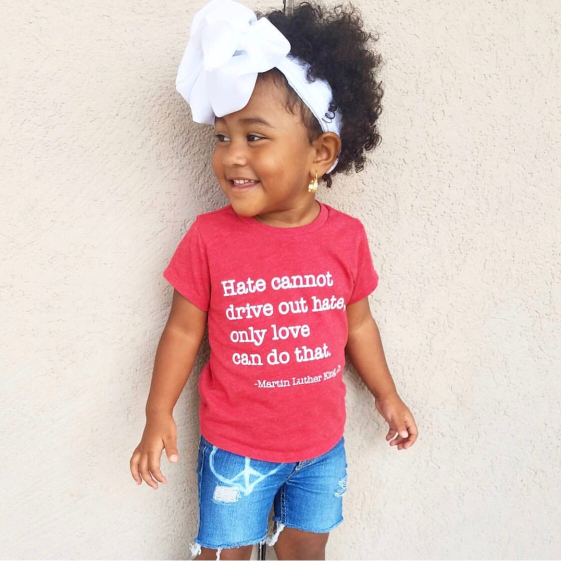 A young girl with curly hair and a large white bow smiles warmly in denim shorts and the MLK Love Tee, standing against a light-colored wall.
