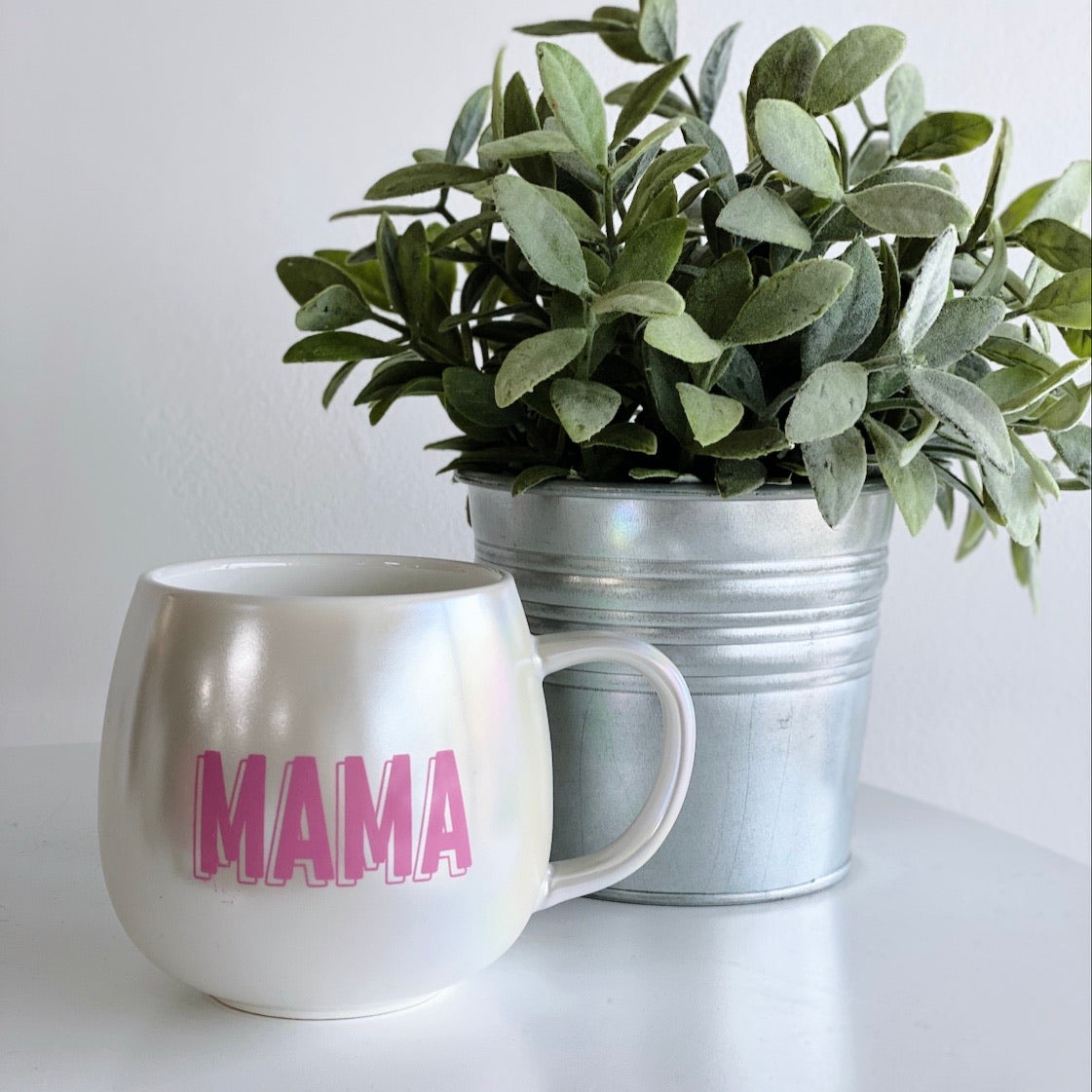 The MAMA Pearl • Ceramic Mug, featuring pink lettering on white ceramic, sits beside a green potted plant in a metallic bucket. HAND-WASH ONLY, this mug adds charm to any space.