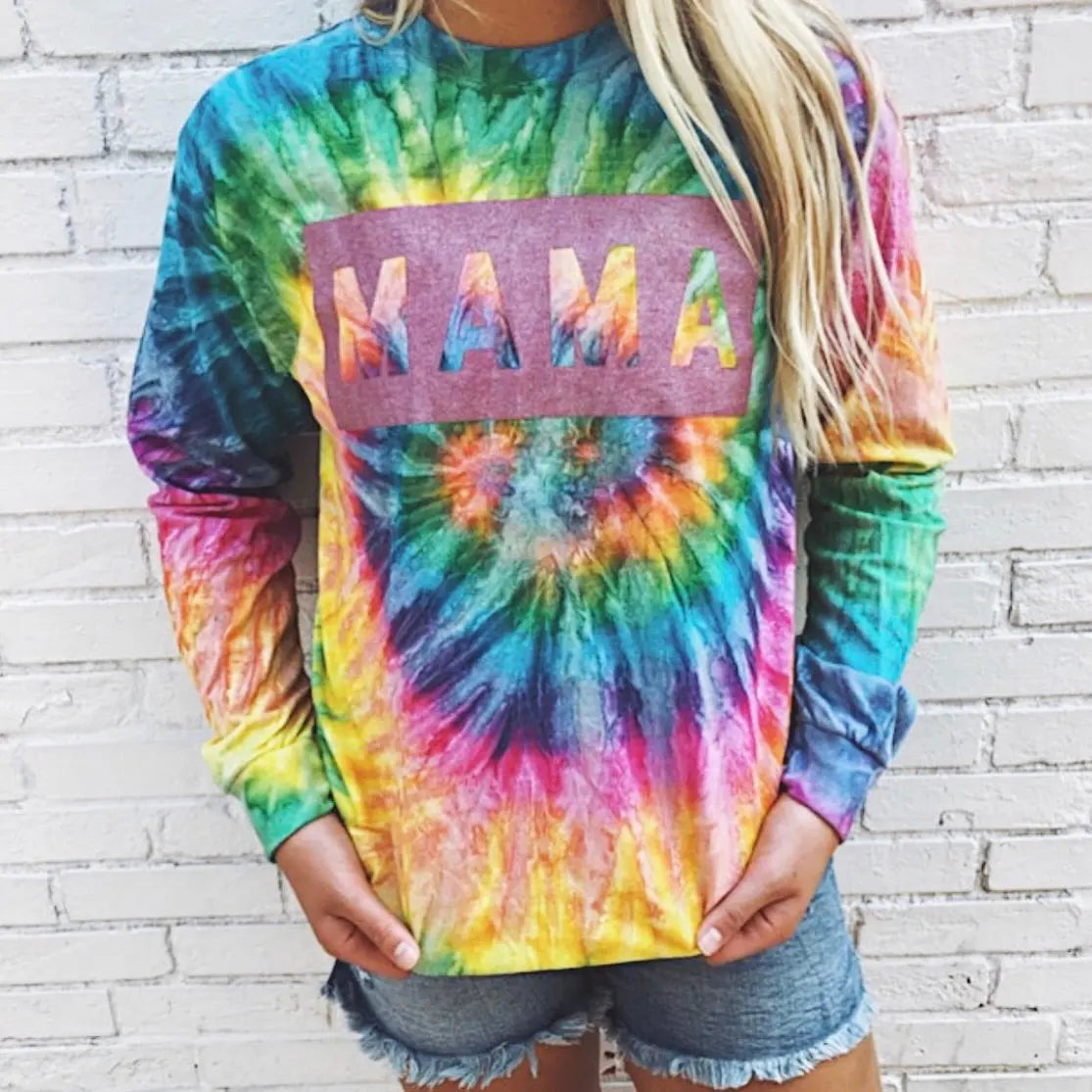 Someone wears the MAMA Box • Longsleeve Tie-Dye shirt, featuring a pink shimmer MAMA design, paired with denim shorts in front of a white brick wall.