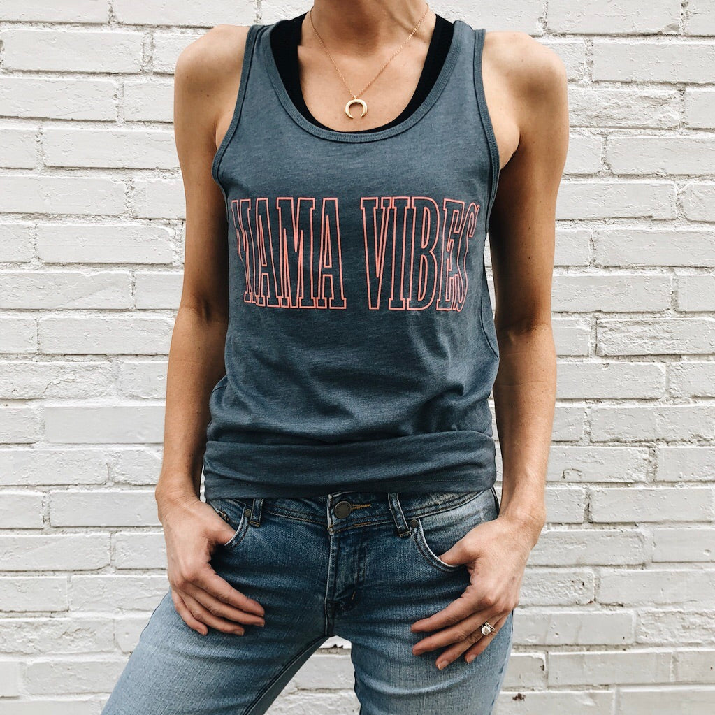 Someone stands against a white brick wall, wearing the MAMA VIBES • Unisex Tank Top in sleeveless gray with bold red lettering, layered over a black undershirt, paired with light blue jeans and a gold necklace. Their face is not shown.