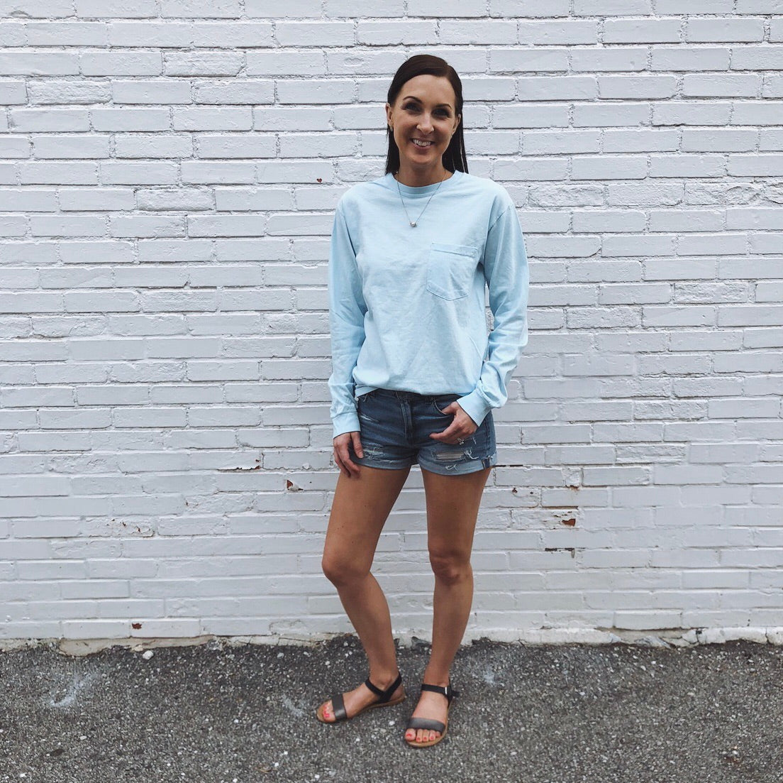 A woman smiles in front of a white brick wall, wearing the Motherhood is Cool Back Print • Long Sleeve shirt in light blue with denim shorts and black sandals, one hand in her pocket on asphalt.