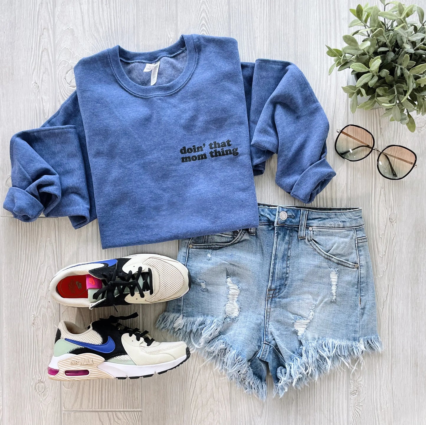 The Mom Thing • Heather Blue Pullover is styled with denim cutoff shorts, black and white sneakers, sunglasses, and a green plant on a light wooden surface.