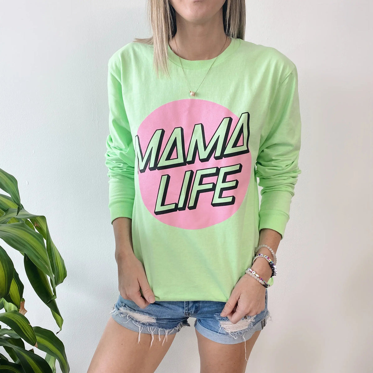 A person wears the Skater Long Sleeve • Mint, a soft pre-washed shirt with a bold pink circle and MAMA LIFE text, paired with denim shorts. A leafy plant appears on the left side of the image.