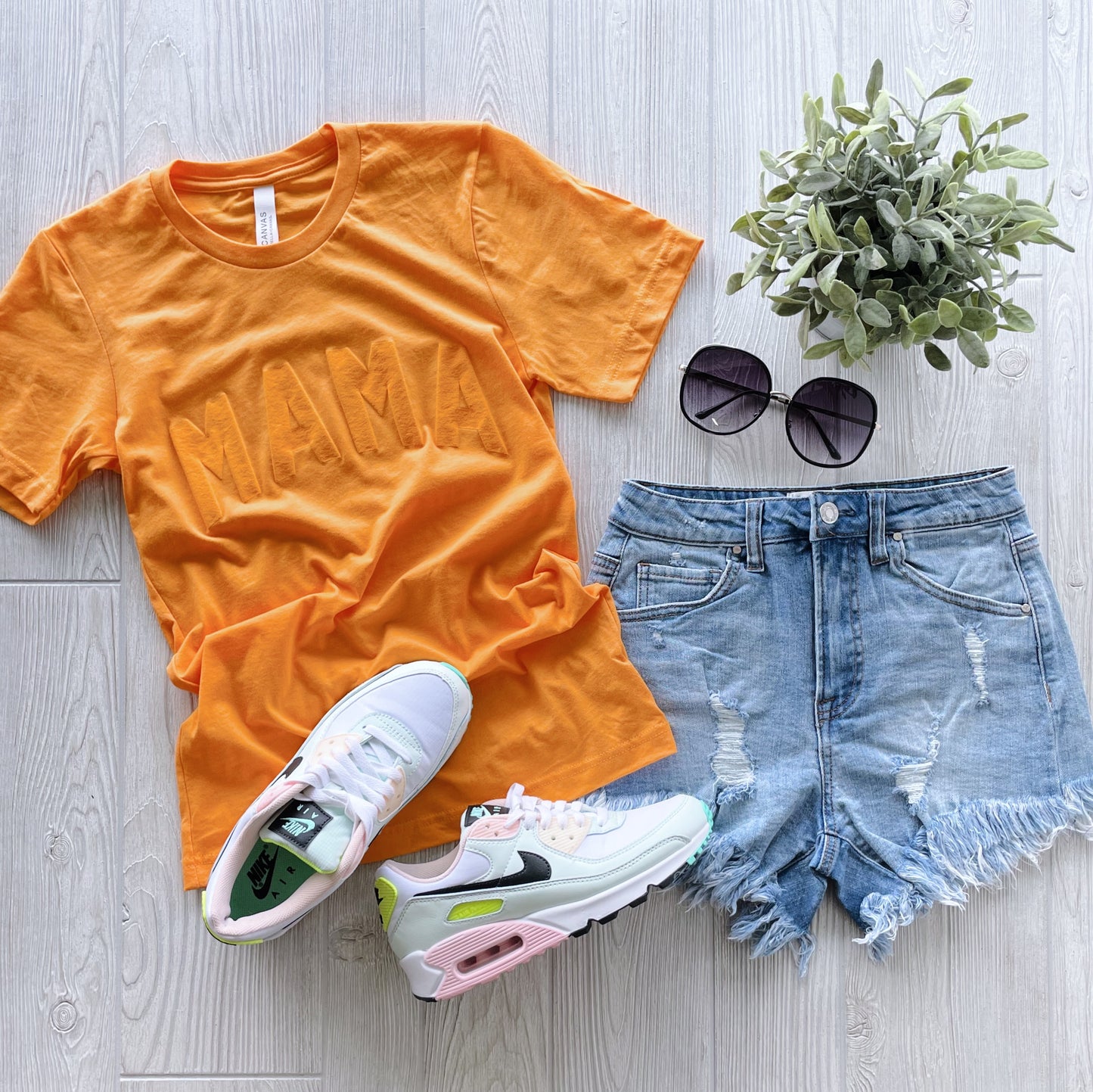 Flat lay of the MAMA • Mango Tonal Tee in a bright orange unisex fit, styled with distressed denim shorts, white Nike sneakers (pink and green accents), black sunglasses, and a small potted plant on light wood.