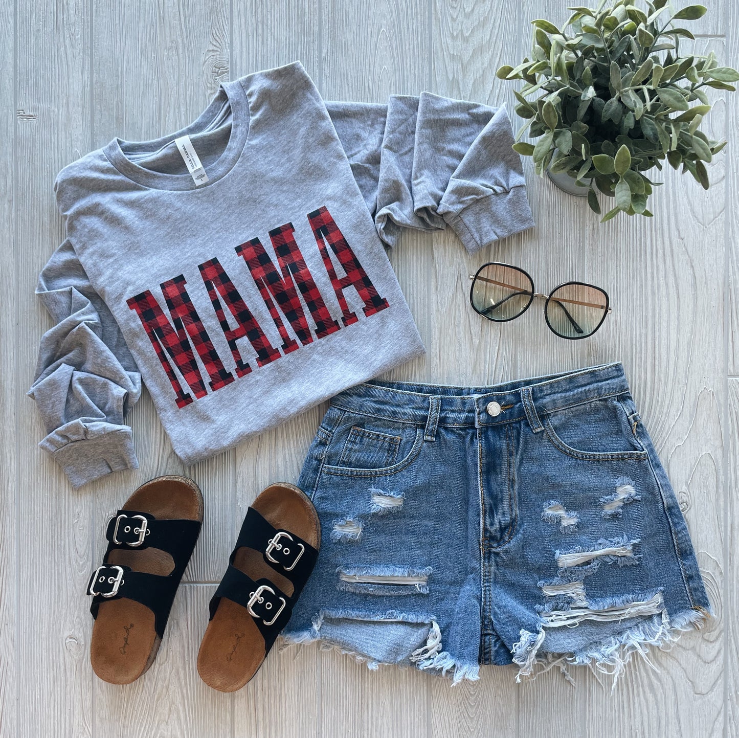 The MAMA Plaid • Long Sleeved, in gray with a buffalo plaid MAMA print, is styled with distressed denim shorts, black sandals, sunglasses, and a small potted plant on a light wood surface.