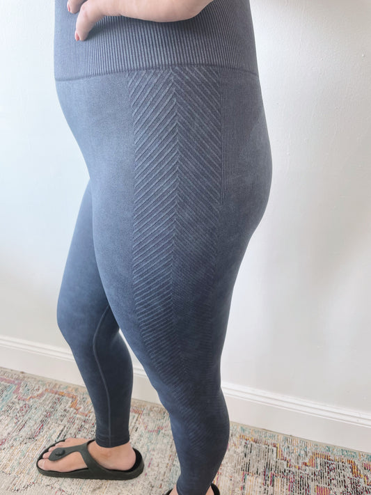 A person wearing Charley Mineral Wash Leggings • 8016 with tummy control and black sandals stands on a colorful, patterned rug against a white wall. Only the lower half of their body is visible.