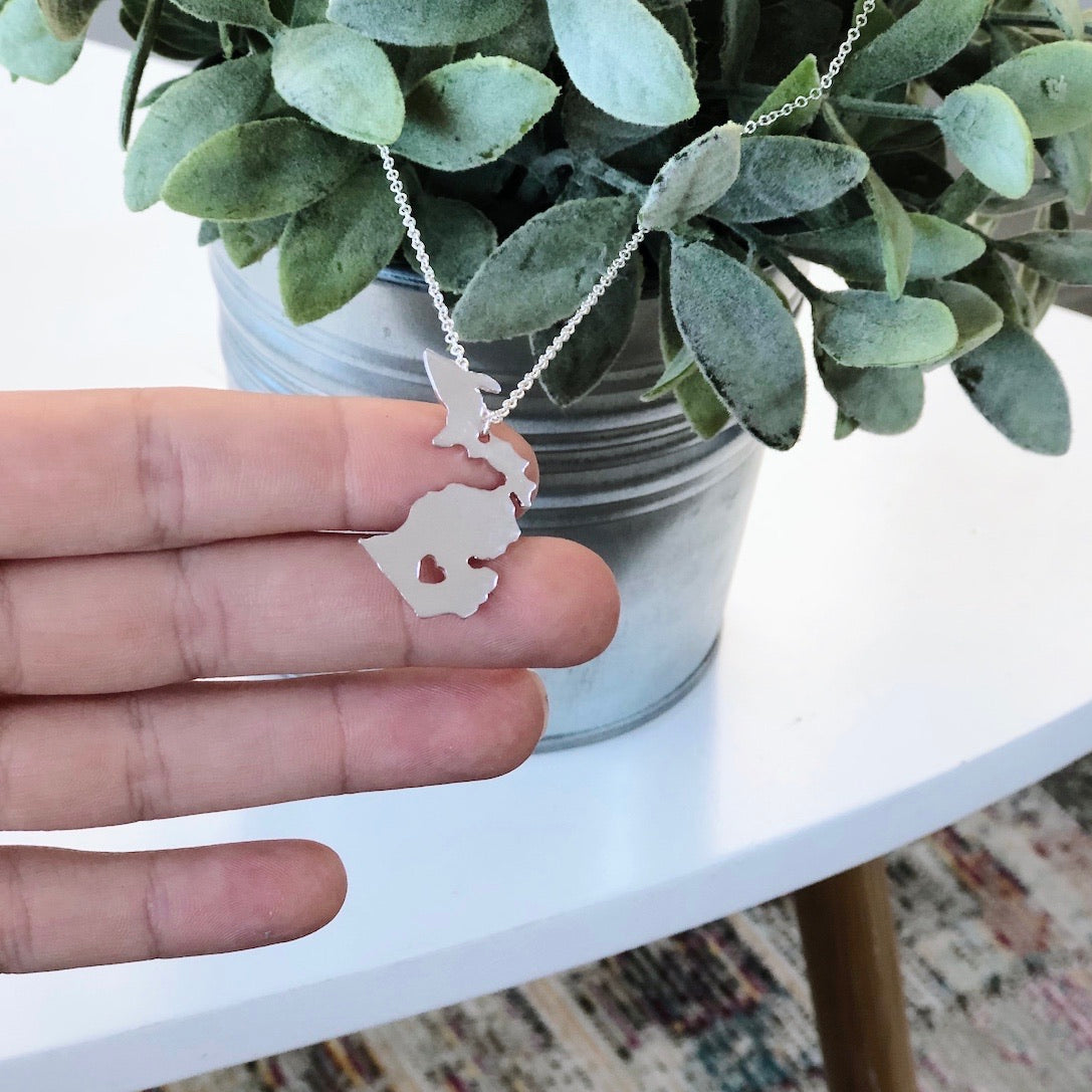 A hand holds a Michigan Necklace • Silver or Gold! with an adjustable clasp, featuring a Michigan-shaped pendant, in front of a potted plant on a white table.