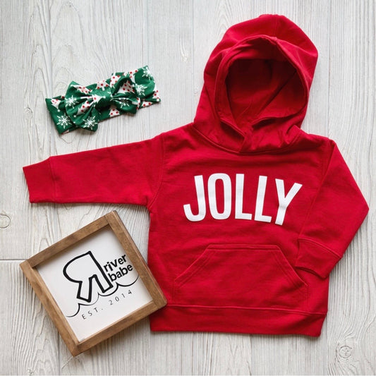 The Jolly Hoodie • Kids in red with a white “JOLLY” print, paired with a festive green headband and a River Babe Est. 2014 logo frame, displayed on light wood—a perfect setup for your jolly little one.