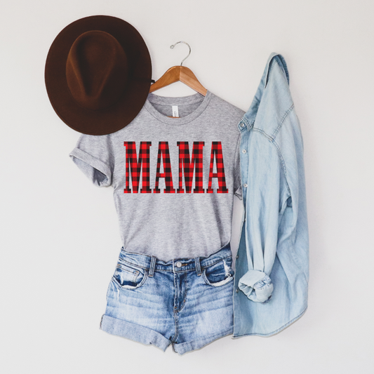 The MAMA • Buffalo Plaid tee in gray features a bold red buffalo plaid print and is shown on a hanger with a brown hat, light denim long-sleeve shirt, and blue denim shorts, all arranged against a white wall.