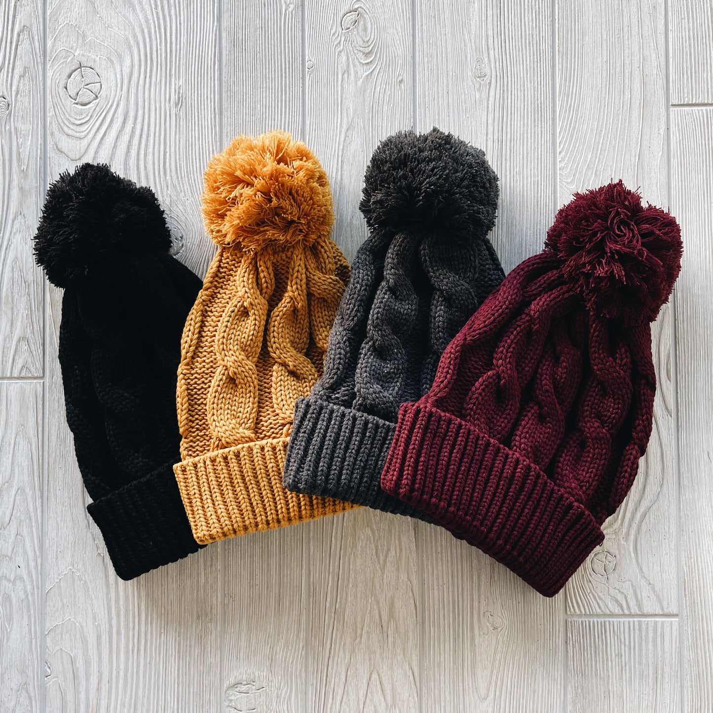Four Cable Knit Pom Beanie • Adult hats in black, mustard yellow, dark gray, and maroon are laid side by side on a light wooden surface.