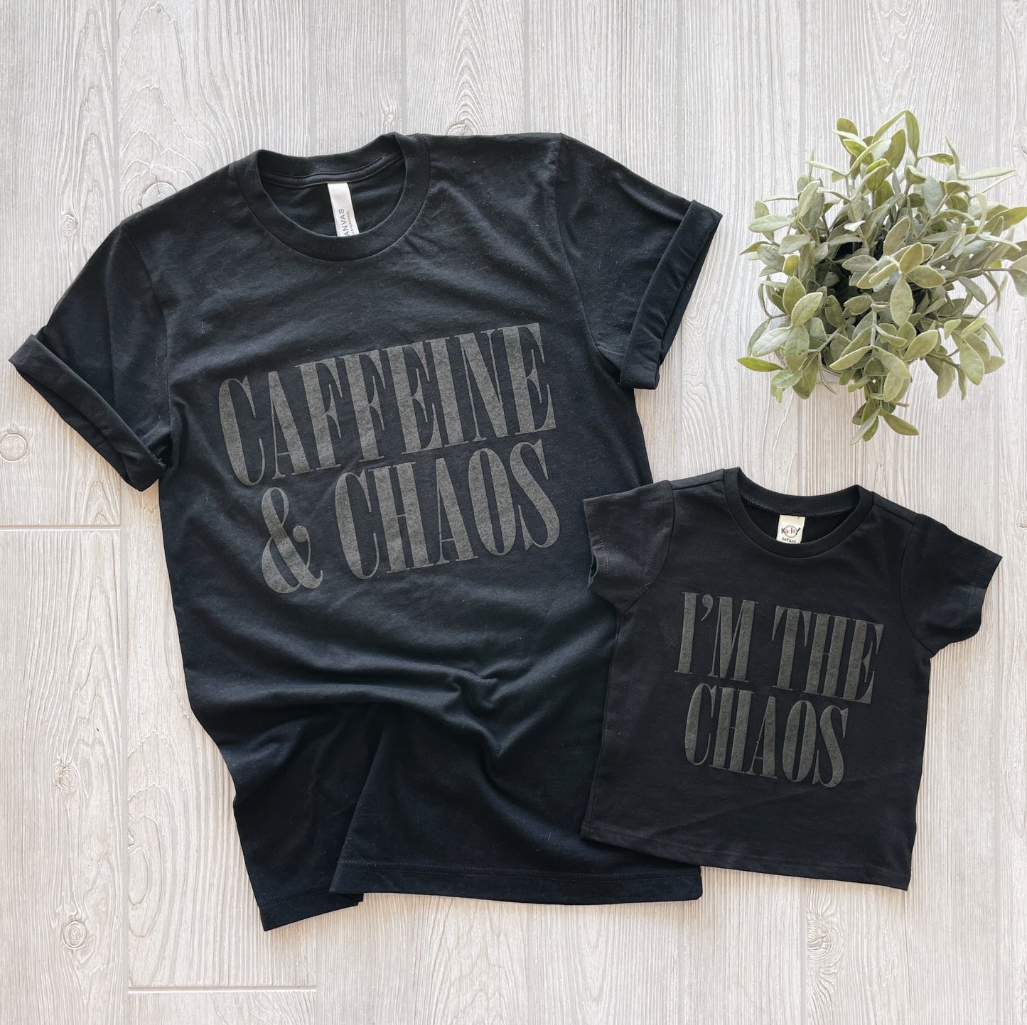 Two black super-soft shirts on a light wooden floor; the larger reads CAFFEINE & CHAOS, and the smaller—CHAOS • Kids Tee, perfect for youth sizes—reads IM THE CHAOS. A small green potted plant finishes off this fun set.