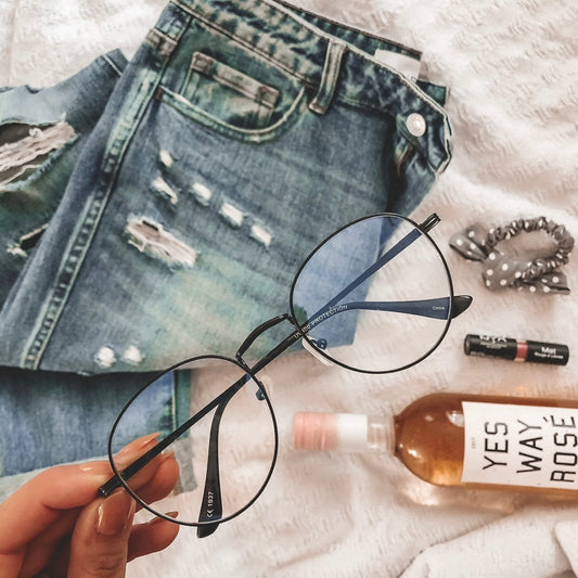 A hand holds Demi Blue Light Glasses (available in more colors) over ripped blue jeans, a scrunchie, lip balm, YES WAY ROSÉ wine, and pale pink lipstick—perfect for reducing screen time headaches.