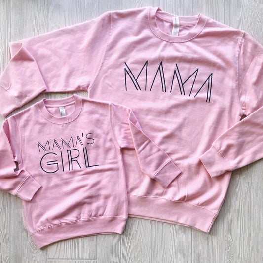 Two light pink unisex fit Mamas Girl Pullover sweatshirts on a light wooden floor. The larger features MAMA in bold, modern letters, while the smaller reads MAMAS GIRL, both in matching black lettering.
