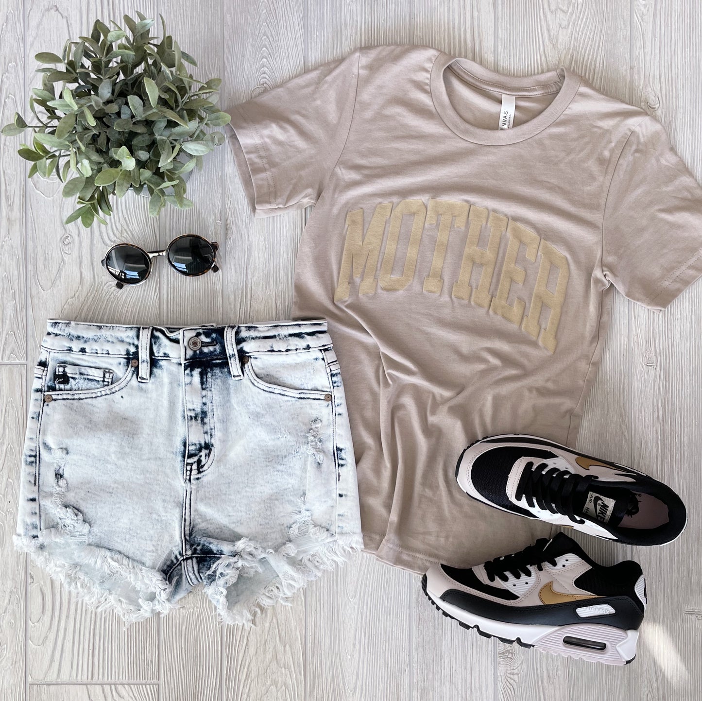 Flat lay of a casual outfit featuring the MOTHER • Tan Tonal Tee, distressed denim shorts, black and white Nike sneakers with gold accents, round sunglasses, and a small potted plant on a light wood background.
