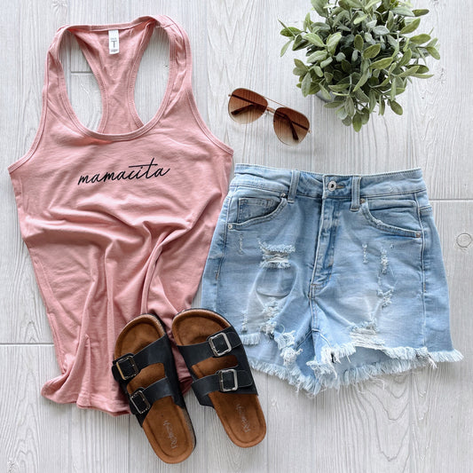 Flat lay of a casual summer outfit: Mamacita • Mauve Racerback tank, distressed light denim shorts, brown sandals, brown sunglasses, and a small green potted plant on a light wood floor.