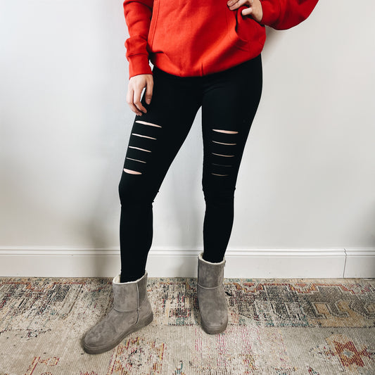 A person stands on a patterned rug, wearing a red hoodie, gray suede boots, and New Shredded Leggings featuring laser-shred distress details. The photo is cropped at the shoulders, so their face is not visible.